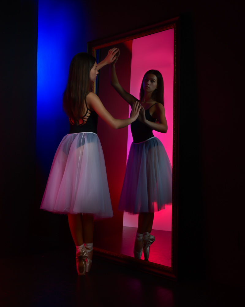 ballerina in fifth looking into mirror with blue and red reflections in background