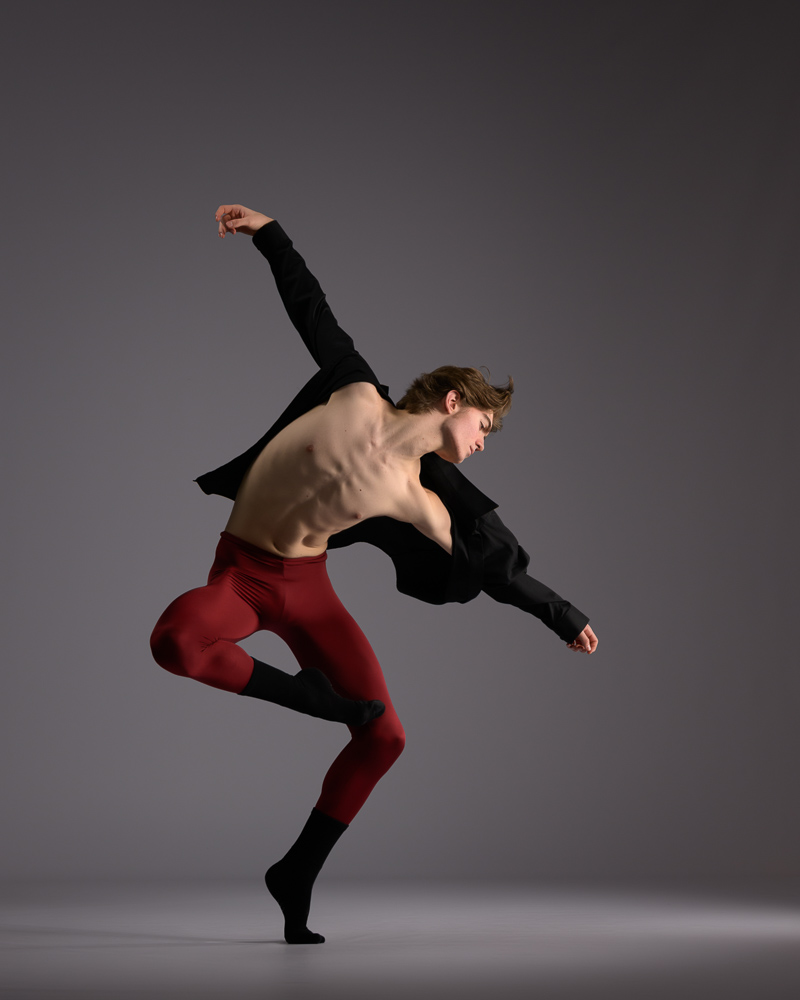 Male dancer in off-balance passe in plie wearing dark red tights and a unbuttoned black shirt.