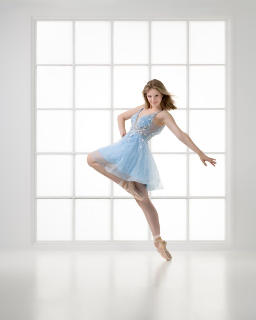 High School ballet dancers in blue dress in fall-over passe in white room with large window.