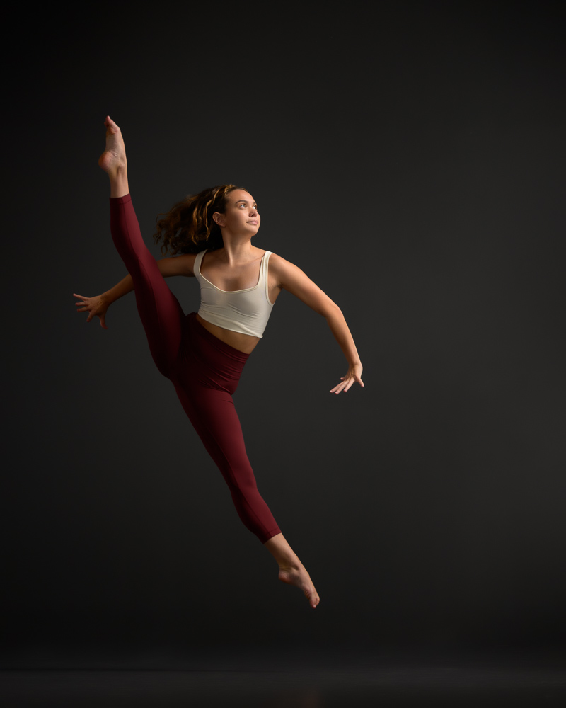 Contemporary female dancer in a straddle jump looking off to the right side with hand flexed inward.