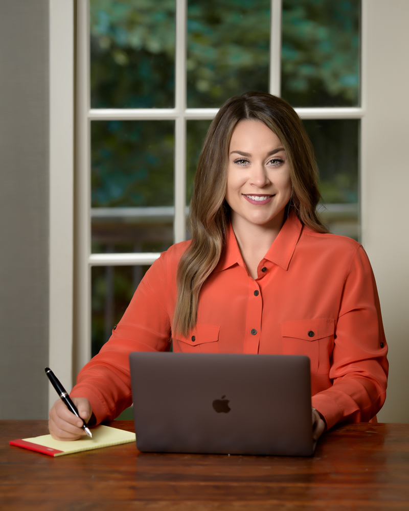 Professional Branding Headshoot of a woman at a desk working on her laptop and taking notes, looking up to the camera with a pleasant smile. Window with trees in the background.