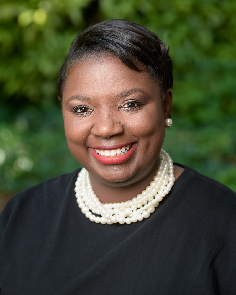 Professional Headshot taken outdoors with a lot of greenery in the background of a black woman with a big smile wearing white pearls