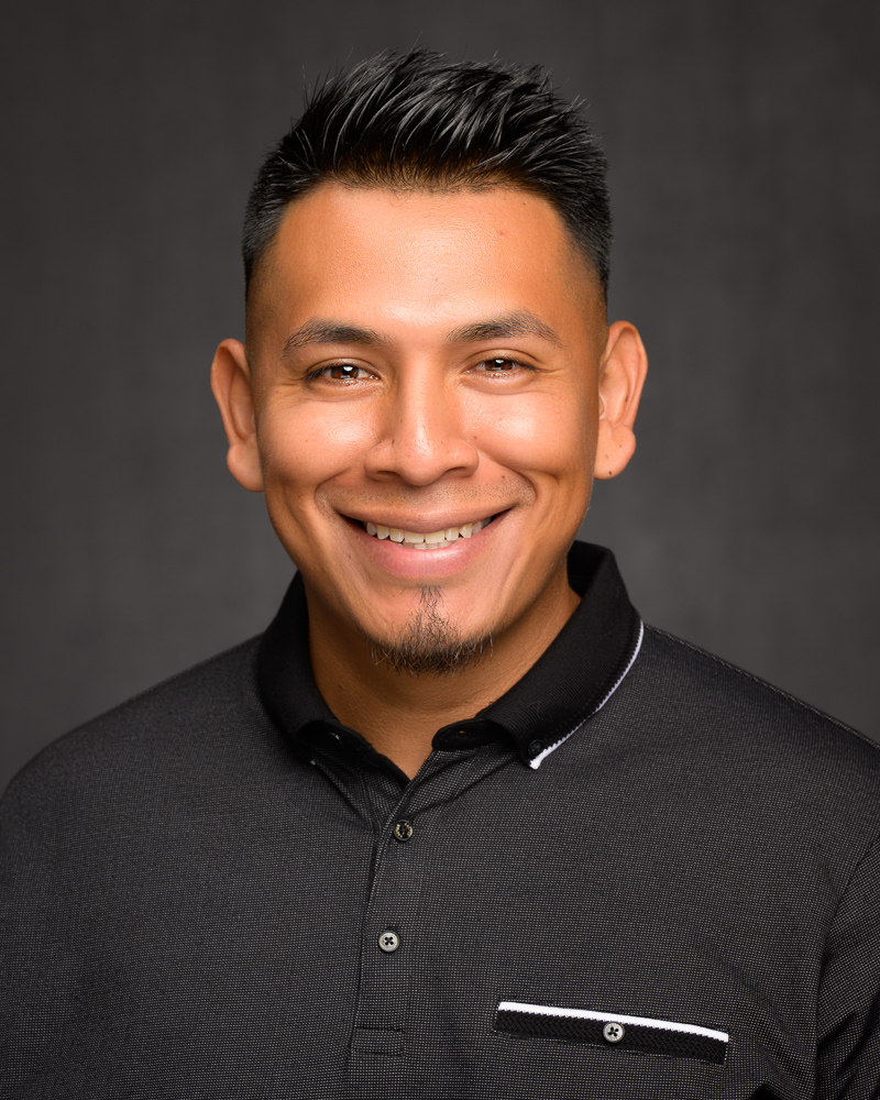 A professional headshot of a latino man with an awesome smile, spiky hair and a goatee wearing a black polo shirt.