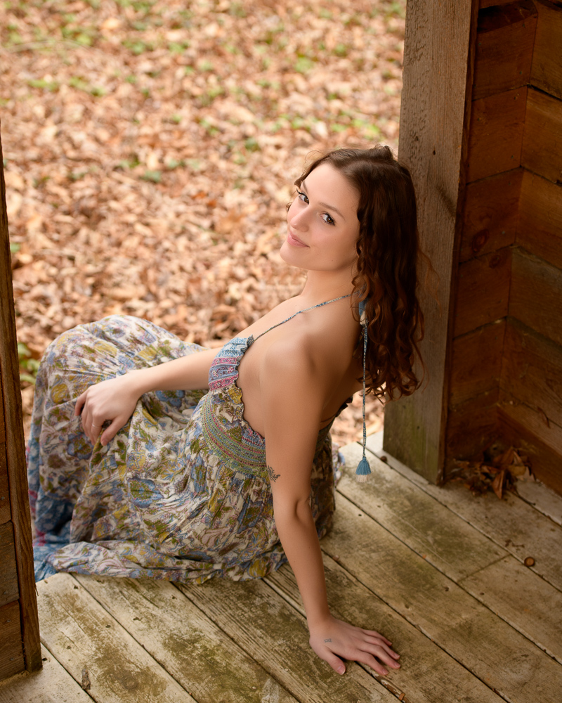 High School senior girl sitting on cabin steps looking back over her shoulder towards the camera with a soft smile.