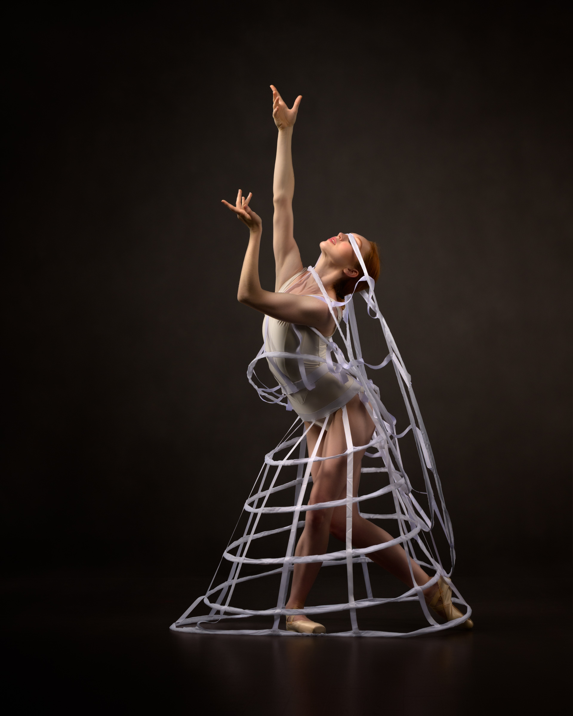 Female ballerina using white cage skirts. One is wrapped around her torso, blind-folding her as she reached skyward.