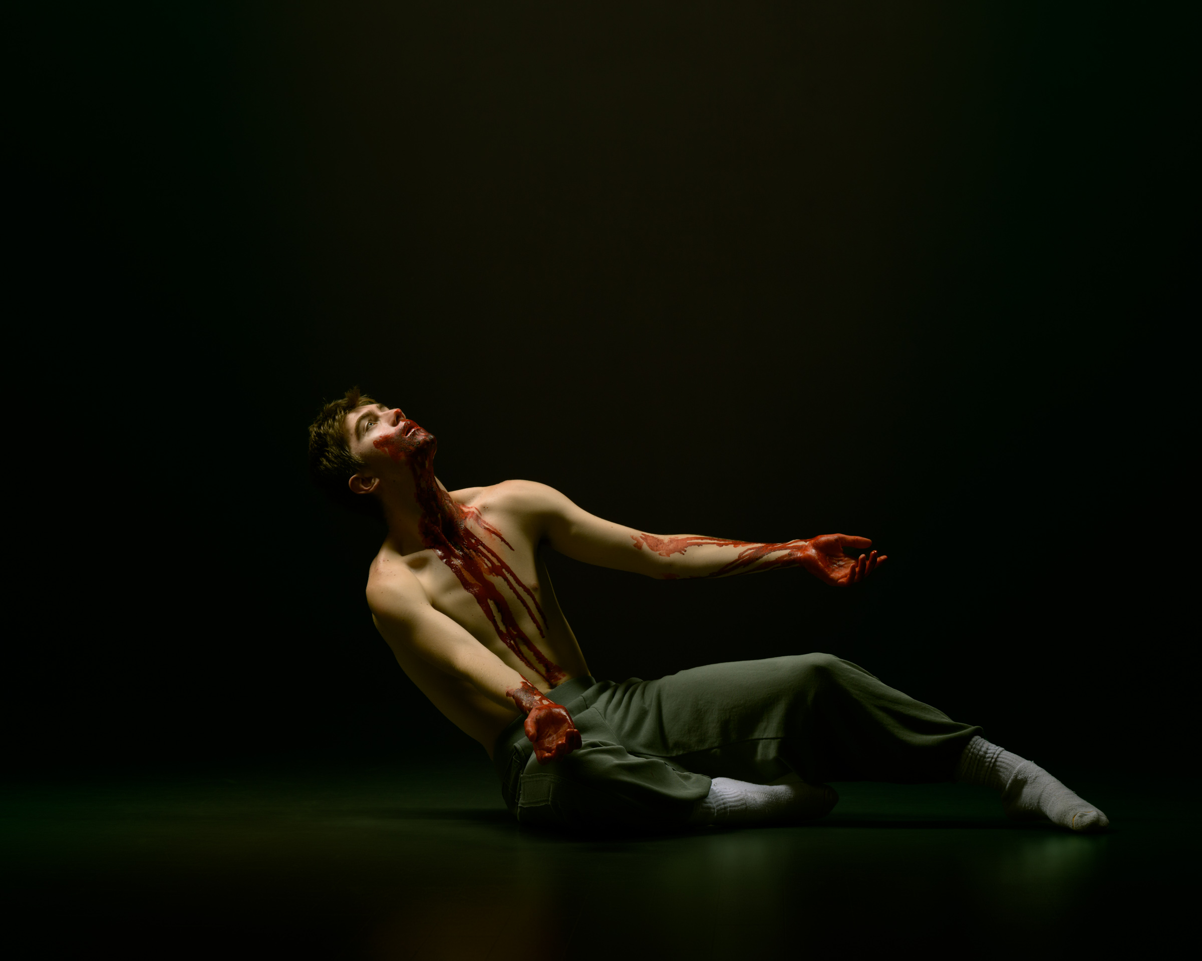 A man begins to fall back. Using fake blood, his hands and torso are covered as he looks up in wonderment.