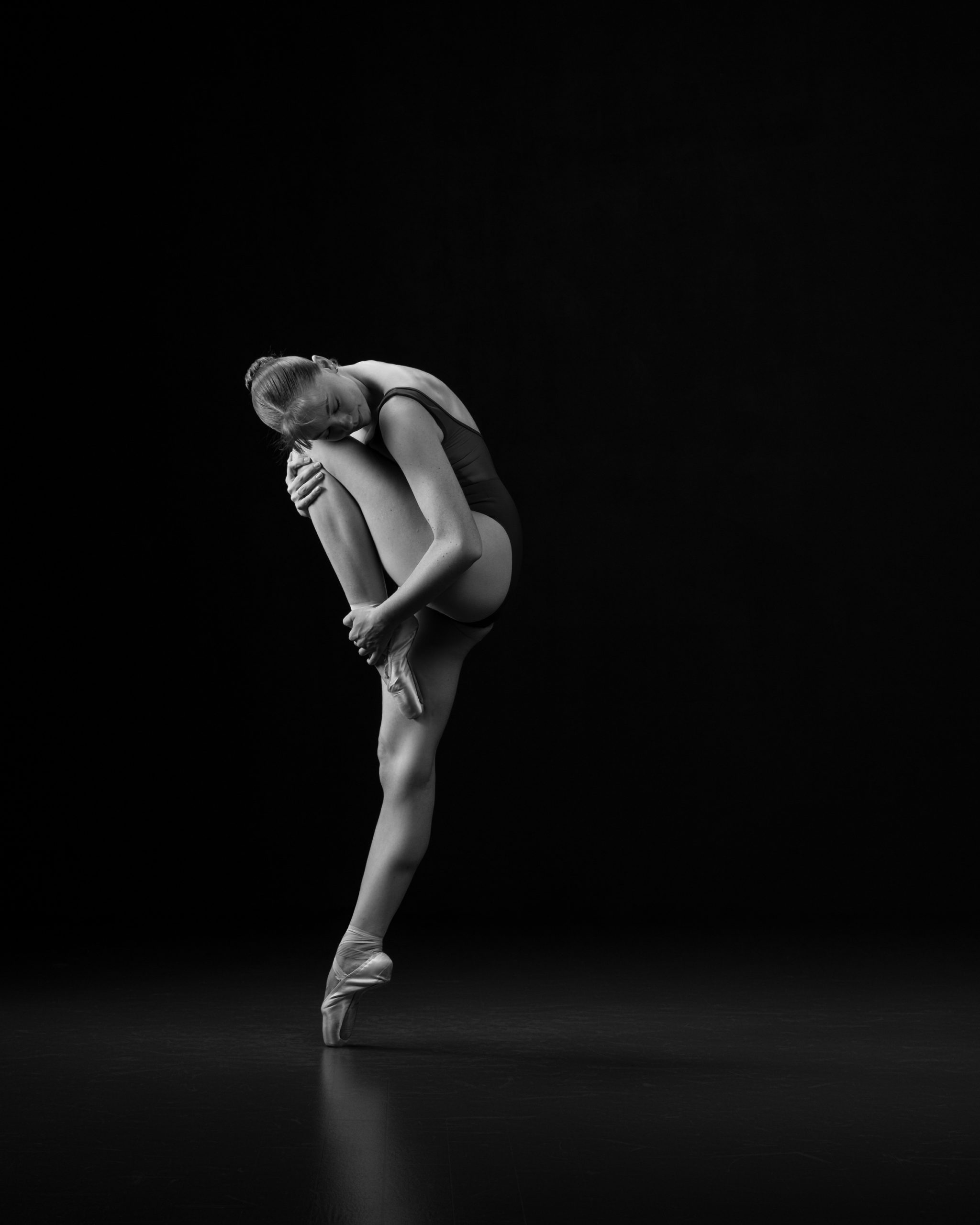 Dancer on pointe holding a very tight passe to her cheek while contracting back off her foot.