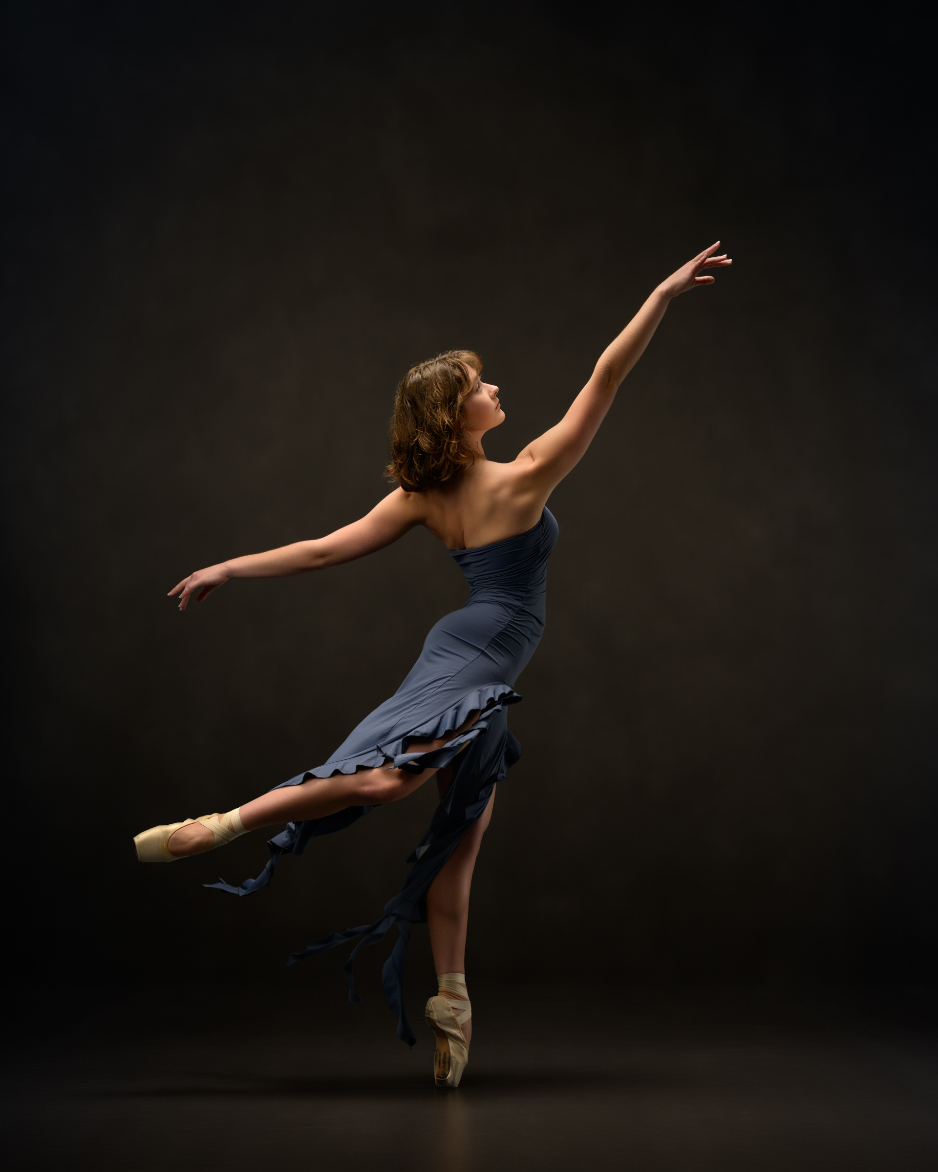 Ballet dancer in blue dress with ruffled fringes in attitude and an long arm lines.
