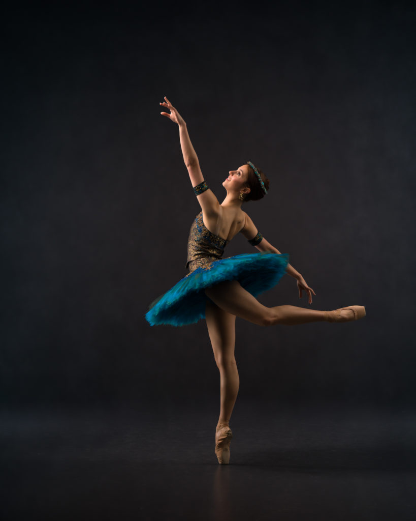 Ballet attitude in blue Pharaoh's Daughter tutu with upward gaze and reach.