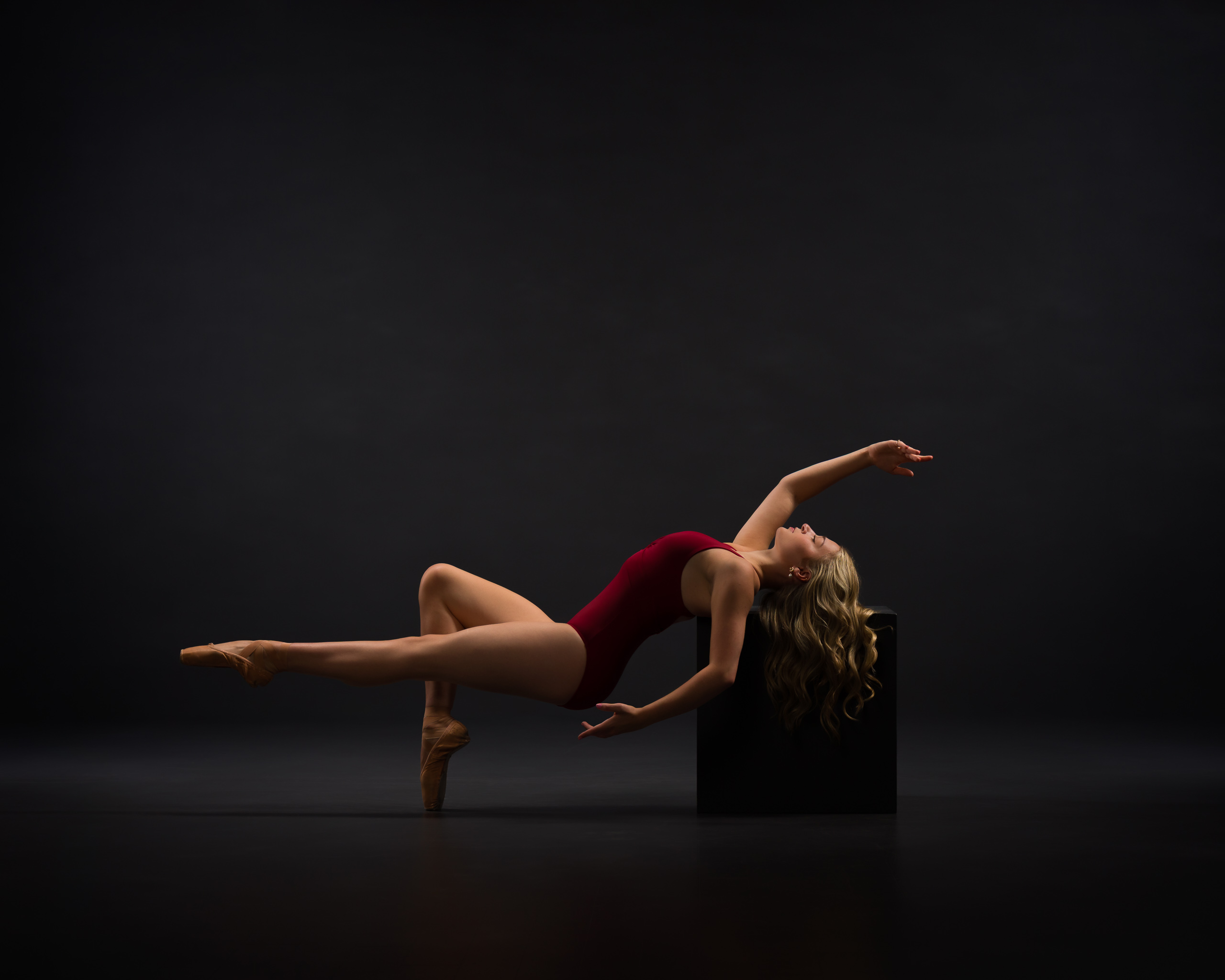 Dancer in a reclined position supported by her head on a box and one foot on pointe. Other leg is straight and parallel to ground. 
