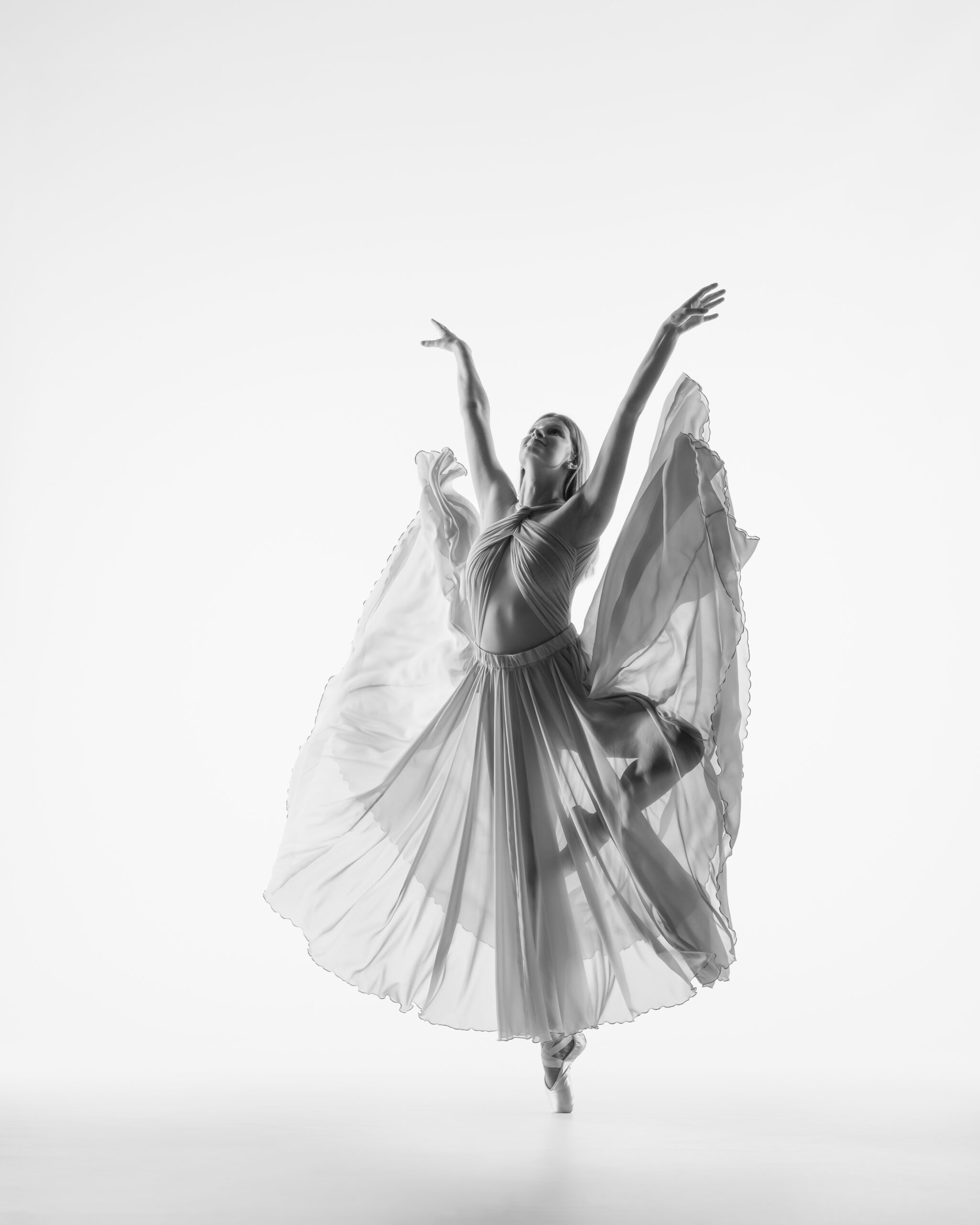 Black and white high key image of HS Senior dance in passe with flowing full skirt and up-stretched arms.