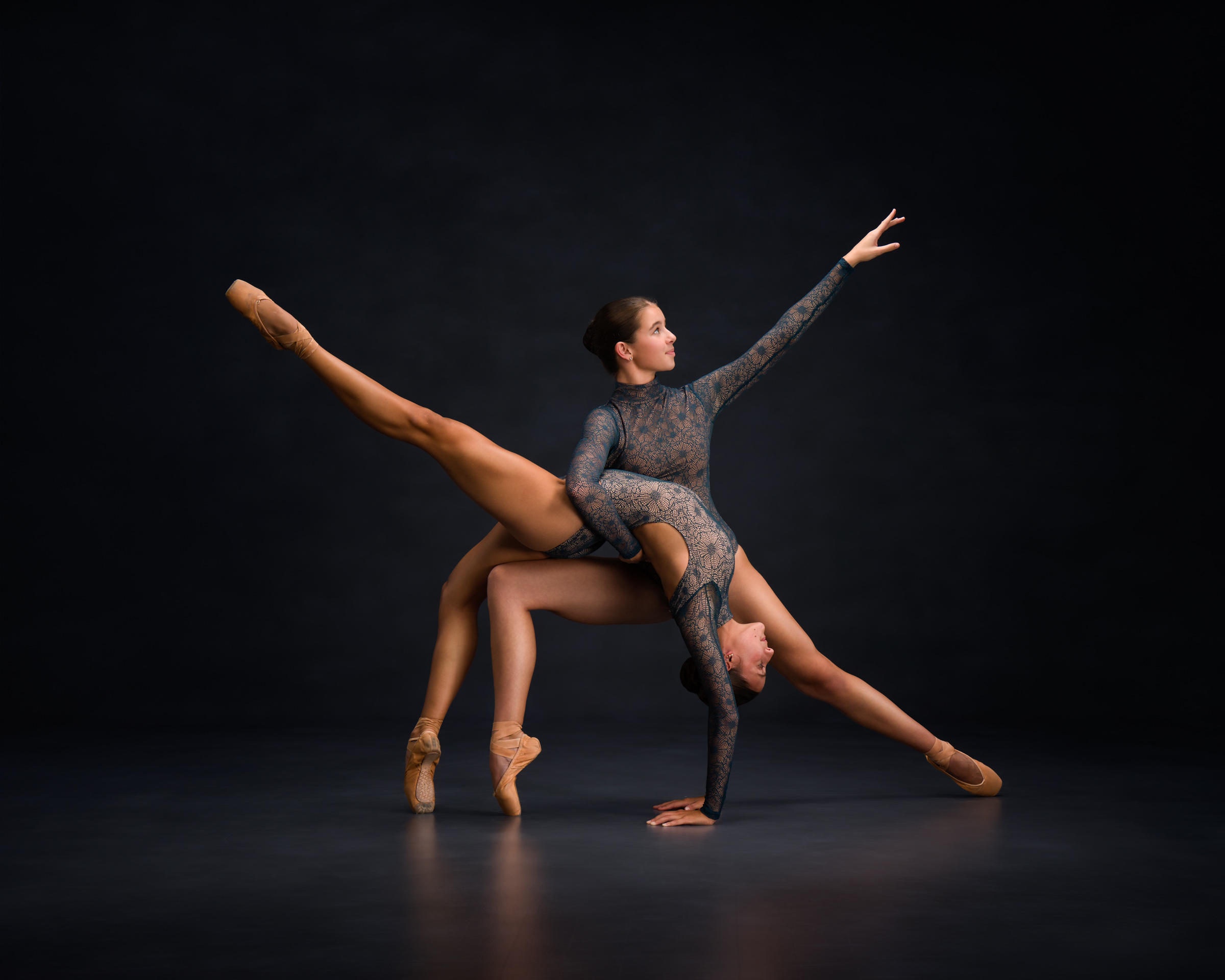 Two dancers in matching leotards one in a lunge and the other in a backbend over the others leg.