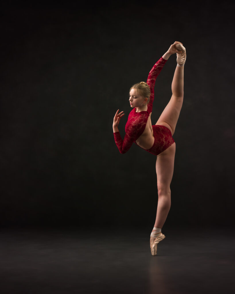 Dancer on pointe in attitude with arm pulling leg high and her gaze downward look past her hand.